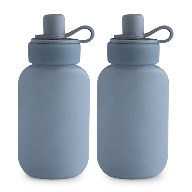 Two blue mushie Silicone Puree Pouches made from BPA-free, food-grade silicone stand side by side, featuring gray caps and looped handles. The front of each pouch displays the embossed mushie brand name.
