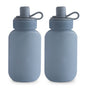 Two blue mushie Silicone Puree Pouches made from BPA-free, food-grade silicone stand side by side, featuring gray caps and looped handles. The front of each pouch displays the embossed mushie brand name.