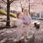 Two young children in light clothes stand on a stone bench beneath blooming cherry blossoms. One holds the mushie Silicone Snack Cup – Easy-Grip Handles, BPA-Free, while the other enjoys a treat amid pink petals scattered on the ground.