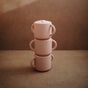 Three pastel pink mushie Silicone Snack Cups with easy-grip handles are stacked against a neutral brown background, softly lit for a warm, minimalistic display ideal for showcasing baby essentials. BPA-free and food-grade safe.