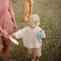 A smiling blond child in a cream sweater and tan shorts holds the mushie Silicone Snack Cup – Easy-Grip Handles, BPA-Free, reaching for a plate from an adult in pink while standing outside on the grass.