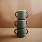 Three mushie Silicone Snack Cup – Easy-Grip Handles, BPA-Free in gray are stacked vertically against a softly lit brown background; the top cup has a sippy lid and the other two are open for convenient snacking.