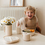 A smiling child in a tan bib sits at a table with fruit and the mushie Silicone Snack Cup – Easy-Grip Handles, BPA-Free; the cozy, light setting with neutral tones is perfect for enjoying baby essentials.