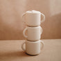 Three matte white mugs with handles are stacked on a beige surface, creating a minimalist look—an ideal match for other baby essentials like the mushie Silicone Snack Cup with easy-grip handles and BPA-free design.