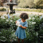 A young girl in a denim dress stands in a garden, pouring tea from the mushie Silicone Tea Play Set – Pretend Play, BPA-Free into a teacup. She is surrounded by lush greenery, blue flowers, and a fountain in the background.