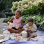 Two young children sit on a picnic blanket outdoors, enjoying a pretend tea party with the mushie Silicone Tea Play Set – Pretend Play, BPA-Free. Green bushes and blooming flowers are in the background.