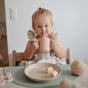 A young blonde child sits at a bright table, smiling and holding the mushie Silicone Training Cup with Straw – BPA-Free, Easy to Hold. In front of her are a plate of banana slices and a cupcake in a minimalistic setting.