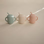 Three mushie Silicone Training Cups with Straws—BPA-free and easy to hold—are shown in mint green, beige, and blush pink on a light surface. Made from food grade silicone, these baby cups feature handles for little hands.