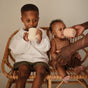 Two young children sit on a wicker bench. The older child in white has a mug, while the baby in brown sips from the mushie Silicone Training Cup with Straw—BPA-free and easy to hold, perfect for baby essentials.