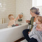 A woman holds her baby in a towel while watching another child play with toy boats and a water pitcher next to the mushie Bath Spout Cover – Soft Silicone, ensuring bath time safety in a white-tiled bathroom with bottles on the tub ledge.