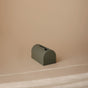 The mushie Bath Spout Cover – Soft Silicone is a small, arch-shaped, matte green silicone piece with grooved lines on one side, sitting on a smooth beige surface against a matching beige background.