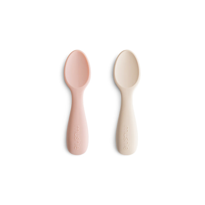 Two mushie Silicone Toddler Starter Spoons in light pink and cream, made from BPA-free, food-grade silicone, lie side by side with mushie embossed on each handle—safe and perfect for encouraging independent feeding.