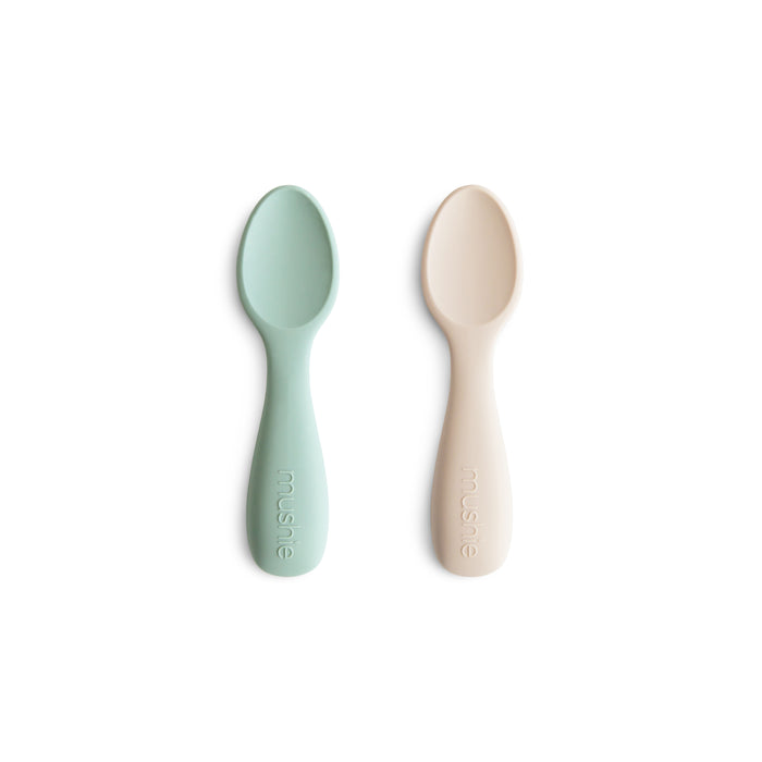 Two mushie Silicone Toddler Starter Spoons, one light green and one beige, lie side by side on a white background. Made from BPA-free, food-grade silicone, each handle is embossed with the brand name mushie.