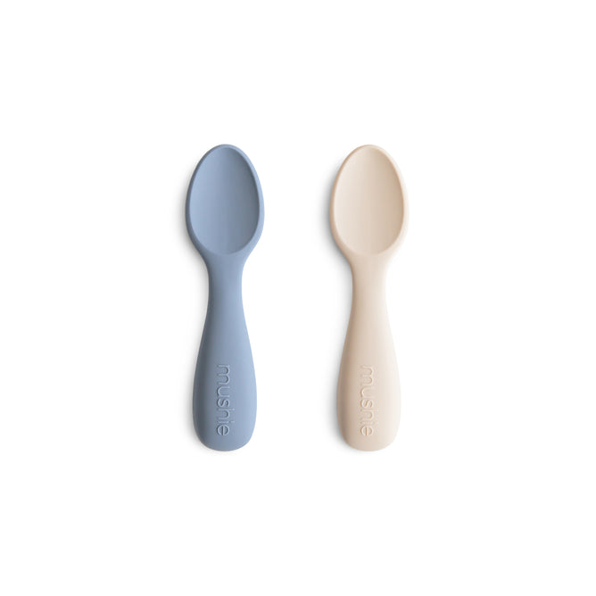 Two mushie Silicone Toddler Starter Spoons—one light blue, one beige—made from BPA-free, food-grade silicone, lie side by side on a white background. Each handle features the mushie logo embossed for a stylish touch.