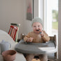 A smiling toddler in a light gray beanie sits in a high chair using mushie Silicone Toddler Starter Spoons—safe, BPA-free, and made from food-grade silicone—with food smeared on their face; a sippy cup and bowl are nearby.