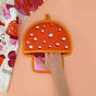 A hand slides into The Neighborgoods Pot Holder- Mushroom, a red, mushroom-shaped pot holder with white spots on a peach surface. A colorful mushroom-themed dish towel peeks from the corner, highlighting this kitchen essentials heat protection.