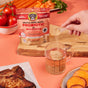 A hand scoops powder from a Brutus on the Go: Probiotics Instant Bone Broth Mega Pack packet into water for canine gut health. Nearby, tomatoes, carrots, herbs, sweet potato slices, and steak sit on a wooden cutting board against a peach background.
