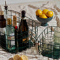 A wire basket holds various bottles and glassware on a checkered cloth. Nearby, a green bowl filled with lemons and a smaller basket with glasses sit on the table, with two lemon halves in front.