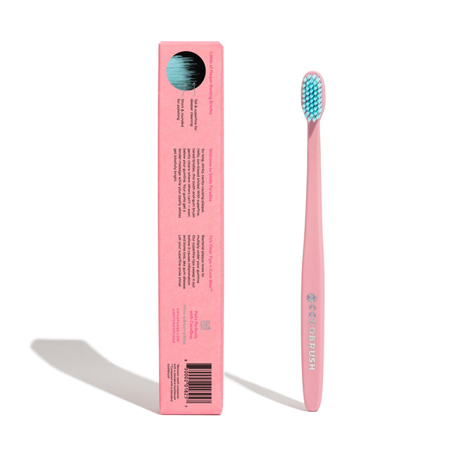 A pink toothbrush stands upright next to its matching pink box, which displays product details and a barcode. The toothbrush has blue and white bristles and both items cast shadows on a white background.
