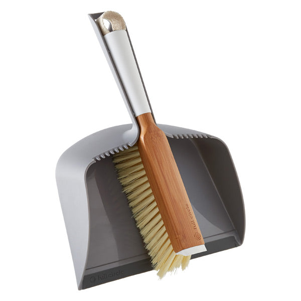 A gray dustpan with a wooden-handled brush resting inside it; the brush has light bristles and fits neatly into the dustpans curved edge.