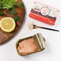 An open tin of Natural Catch Tuna - Yellowfin in Olive Oil is displayed on a white surface with a half lemon, cilantro, a fork with tuna, and its colorful box labeled Natural Catch, rich in Omega-3s.