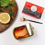 An open tin of Natural Catch Tuna - Spicy Yellowfin Filets in Extra Virgin Olive Oil is displayed with a lemon slice, cilantro, a small fork holding tuna, and the colorful Natural Catch packaging on a white surface.