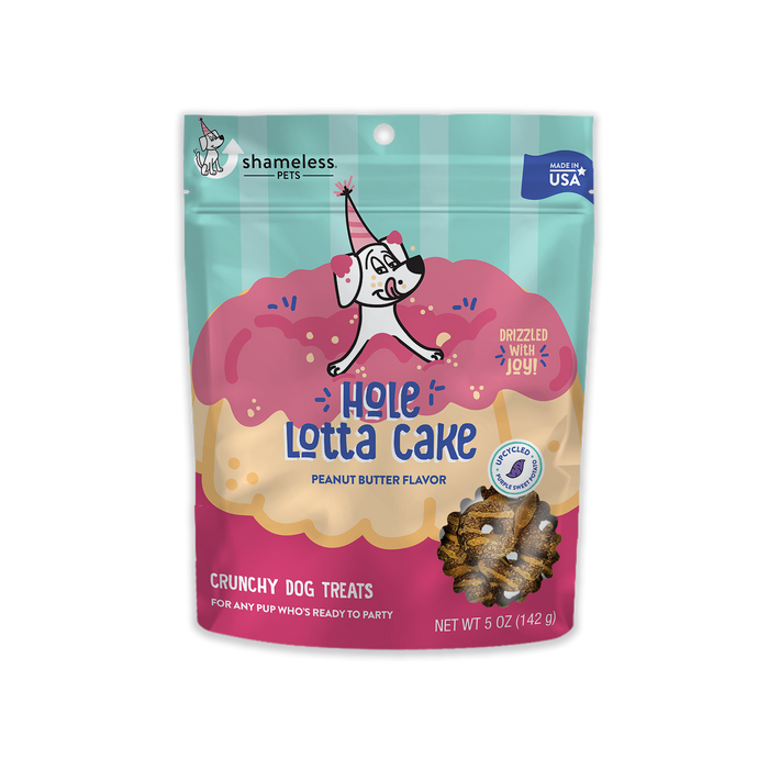 A colorful bag of Shameless Pets “Hole Lotta Cake” peanut butter-flavored crunchy dog treats, featuring an illustrated dog in a party hat on a pink and tan frosted cake. The bag is 5 oz and labeled Made in USA.
