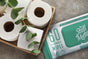 A basket of toilet paper rolls with a eucalyptus branch on top rests on a stone surface. Next to it is a pack of Stall Mates Flushable Wipes, which feature Hydraspun Dispersible technology and are hypoallergenic, as well as alcohol-, paraben-, and fragrance-free.