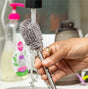 A hand rinses a gray Double-Ended Silicone Cleaning & Bottle Brush from Haakaa under running water, surrounded by soap bubbles. In the background, a dish soap bottle with a pink pump and a brown water bottle complete the scene, highlighting essential hygiene tools.