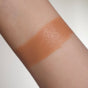 A forearm with a rectangular swatch of medium tan foundation makeup, showing the contrast between the product and natural skin tone. The background is plain and light-colored.