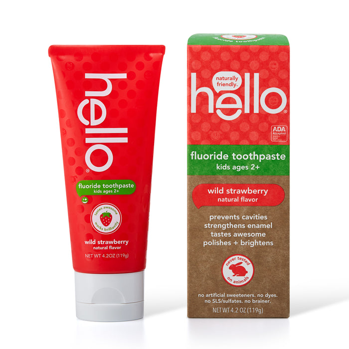 A red tube and matching box of Hello brand fluoride toothpaste for kids, wild strawberry flavor, displayed upright. The box highlights benefits like preventing cavities and strengthening enamel.
