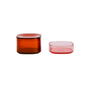 Two transparent containers: one taller, round, and amber-colored with a lid on the left, and one shorter, square, and pink without a lid on the right, both on a white background.