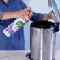 A person in a blue shirt sprays disinfectant into an open stainless steel trash can on a white countertop.