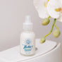 A bottle of Nellies Toilet Spray - Lemongrass with a spray nozzle rests on a white toilet tank beside a blurred white orchid. Its label sports a blue vintage logo and text, emphasizing the green formula and lemongrass scent.