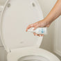 A hand sprays Nellies Toilet Spray - Lemongrass over an open white toilet seat, infusing the clean bathroom with a crisp citrus scent.