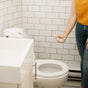 A person in a yellow top and jeans sprays Nellies Toilet Spray - Lemongrass into a white toilet in a bathroom with white tiled walls and a folded towel on the tank.