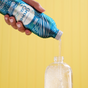 A person pours Grove Co.s Dish Soap Refill - Spring Collection into a clear bottle with a blue and teal pattern against a yellow background. The liquid is clear, and the hand holds the white-capped bottle, showcasing Grove Collaboratives dedication to reducing plastic pollution.