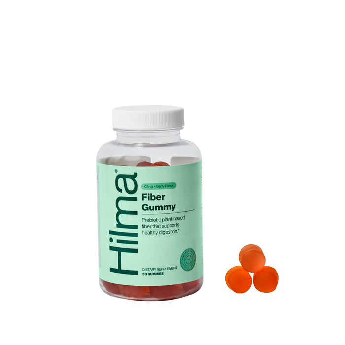 A clear bottle labeled Hilma Fiber Gummy with a green label, containing red-orange gummies. Three gummies are placed outside the bottle, next to it, on a white background.