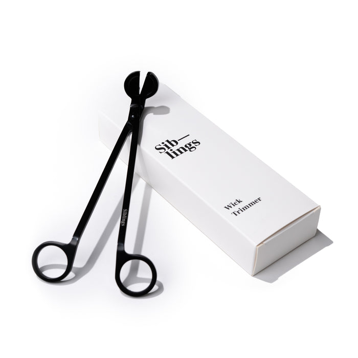 Black wick trimmer with looped handles resting on a white box labeled Siblings Wick Trimmer in black text, all on a white background.