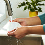 Close-up of hands washing a white dish under running water from a kitchen faucet, with a potted plant in a yellow pot blurred in the background.