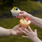 Three hands holding cupcakes with swirled frosting; one cupcake is topped with a blueberry and leaf, and the other with a sliced strawberry and a leaf. The background is outdoors and blurred.