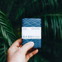A hand holds a blue, patterned Oars + Alps soap bar from the Athlete Kit, designed for deep cleaning and exfoliation, with green leafy plants in the background.