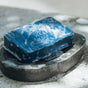A blue bar of soap with white suds sits on a dark dish, part of the Oars + Alps Athlete Kit, and is placed on a light-colored textured surface.