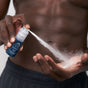 A person sprays a fine mist from a blue Oars + Alps Athlete Kit bottle onto their palm, with their bare torso and dark shorts visible in the background.