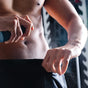 A shirtless person sprays Oars + Alps Skin Saver Body Spray into the waistband of their black athletic shorts, focusing on their torso and hand, with gym equipment blurred in the background.