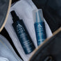 Two Oars + Alps Skin Saver Body Spray bottles, a rinse-free cleanser with Hypochlorous Acid, sit on a white towel in a bag beside a white sneaker.