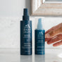 Two blue Oars + Alps Skin Saver Body Spray bottles sit on a white surface. A hand reaches for one. The bottles display white text and a minimalist mountain logo.