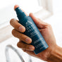 A hand holds a blue bottle of Oars + Alps Skin Saver Face Spray with hypochlorous acid near a sink and blurred faucet—a soothing solution for irritated skin.