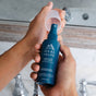 Someone holds a blue bottle of Oars + Alps Skin Saver Face Spray and removes its clear cap near a bathroom sink and mirror.