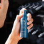A person holds up an Oars + Alps Skin Saver Face Spray bottle with a blurred dark background. The blue bottle highlights hypochlorous acid and hyaluronic acid for soothing irritated skin, and it’s alcohol-free.